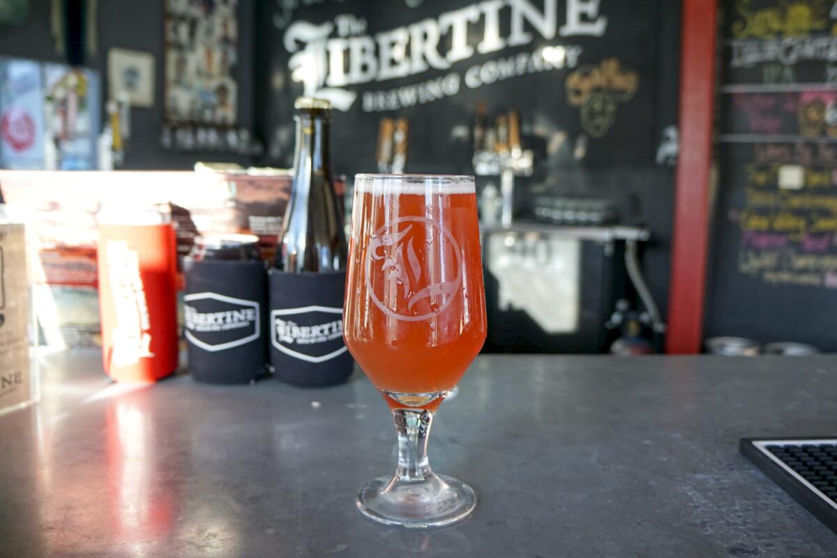 A goblet of reddish beer on a counter in front of a sign that says The Libertine Brewing Company