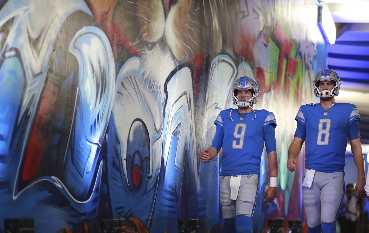 Two Detroit Lions quarterbacks, in uniform, walk past a stadium tunnel mural.