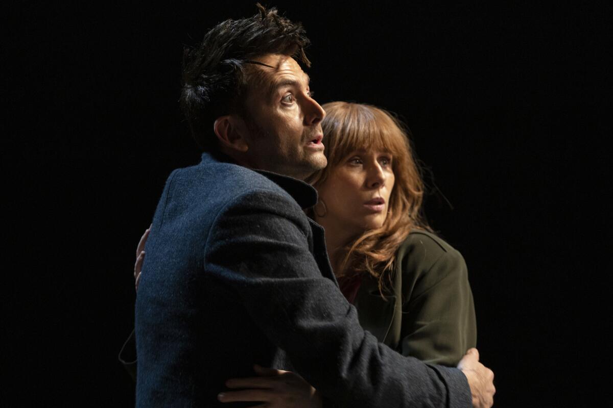 The Doctor, in a dark coat, and Donna Noble, in a green coat, embrace and look upward.