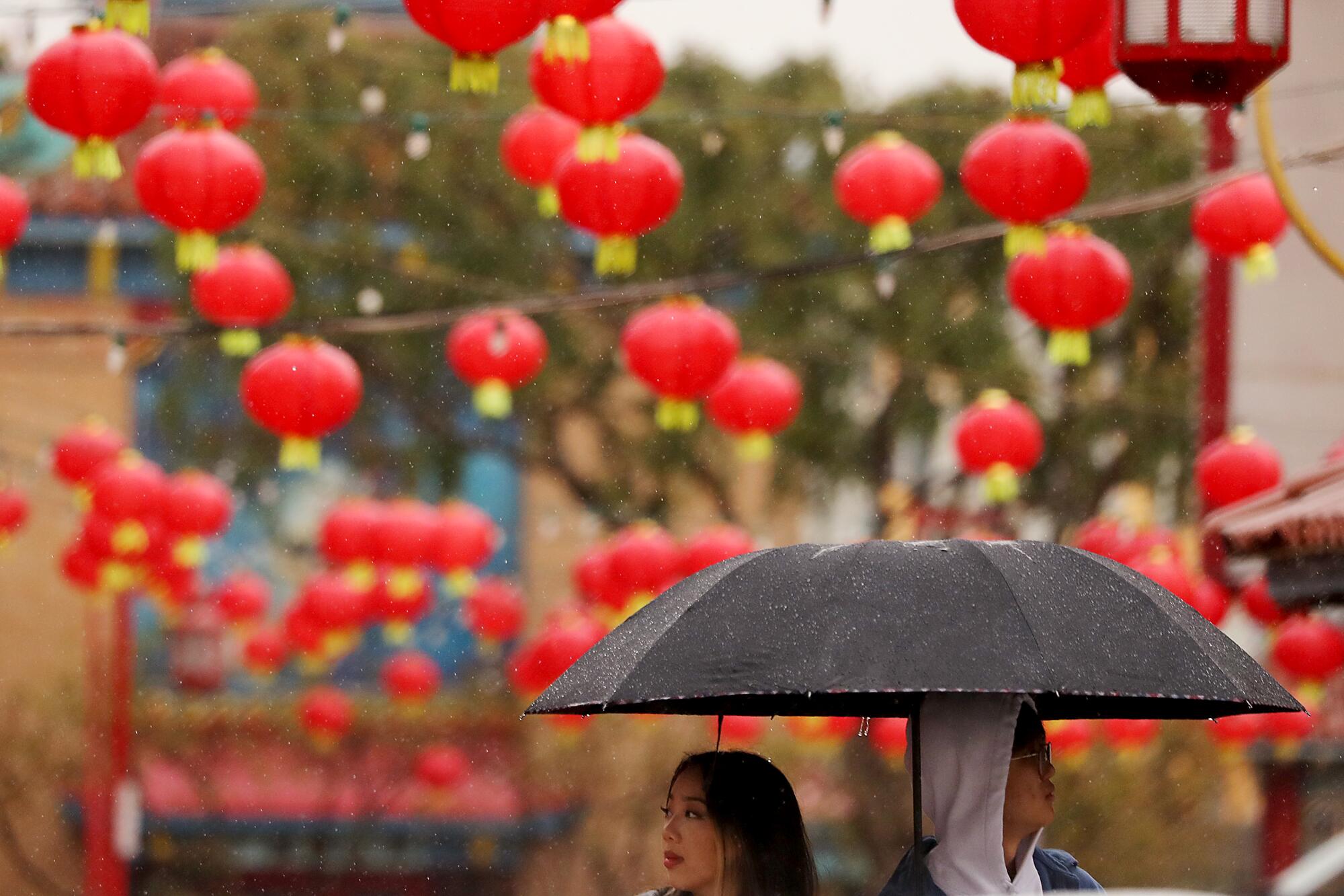 Dozens of red, round Chinese lecterns hang over a plaza near two people under an umbrella.