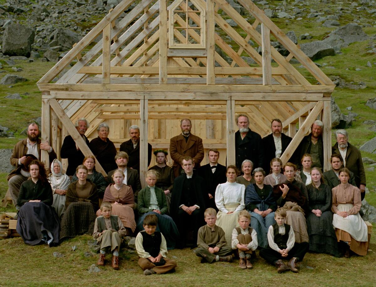 More than two dozen men, women and children in period dress take a group portrait in front of a building under construction.