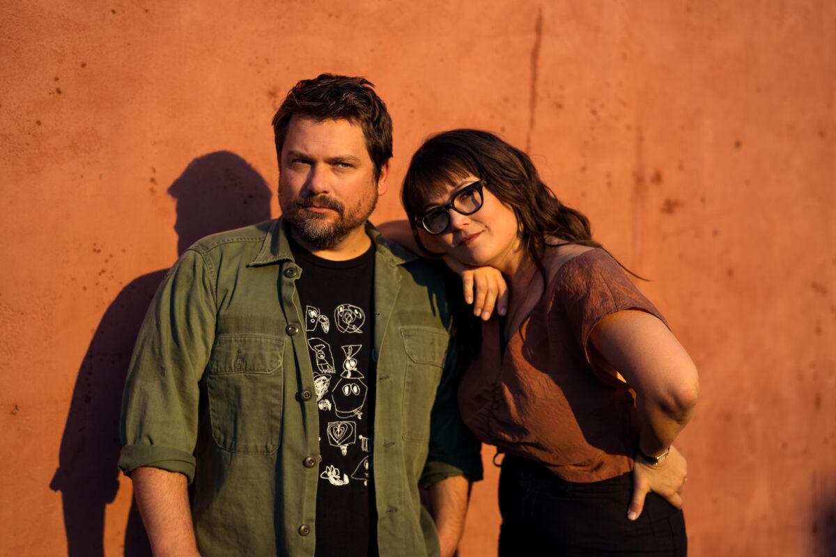 A woman in glasses leans her arm on the shoulder of a bearded man as they pose for a picture against a concrete wall.