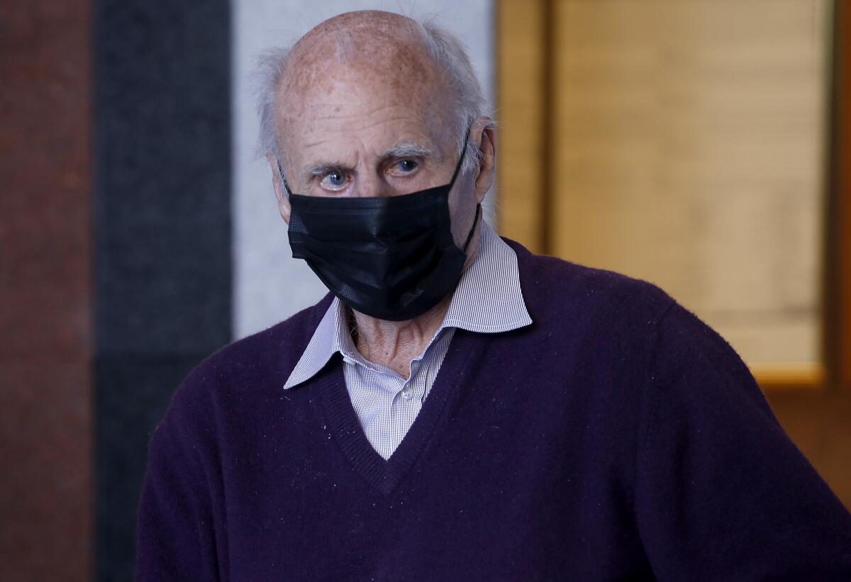 A head-and-shoulder image of an elderly-looking man in black facial mask, purple v-neck sweater and a collar shirt. 