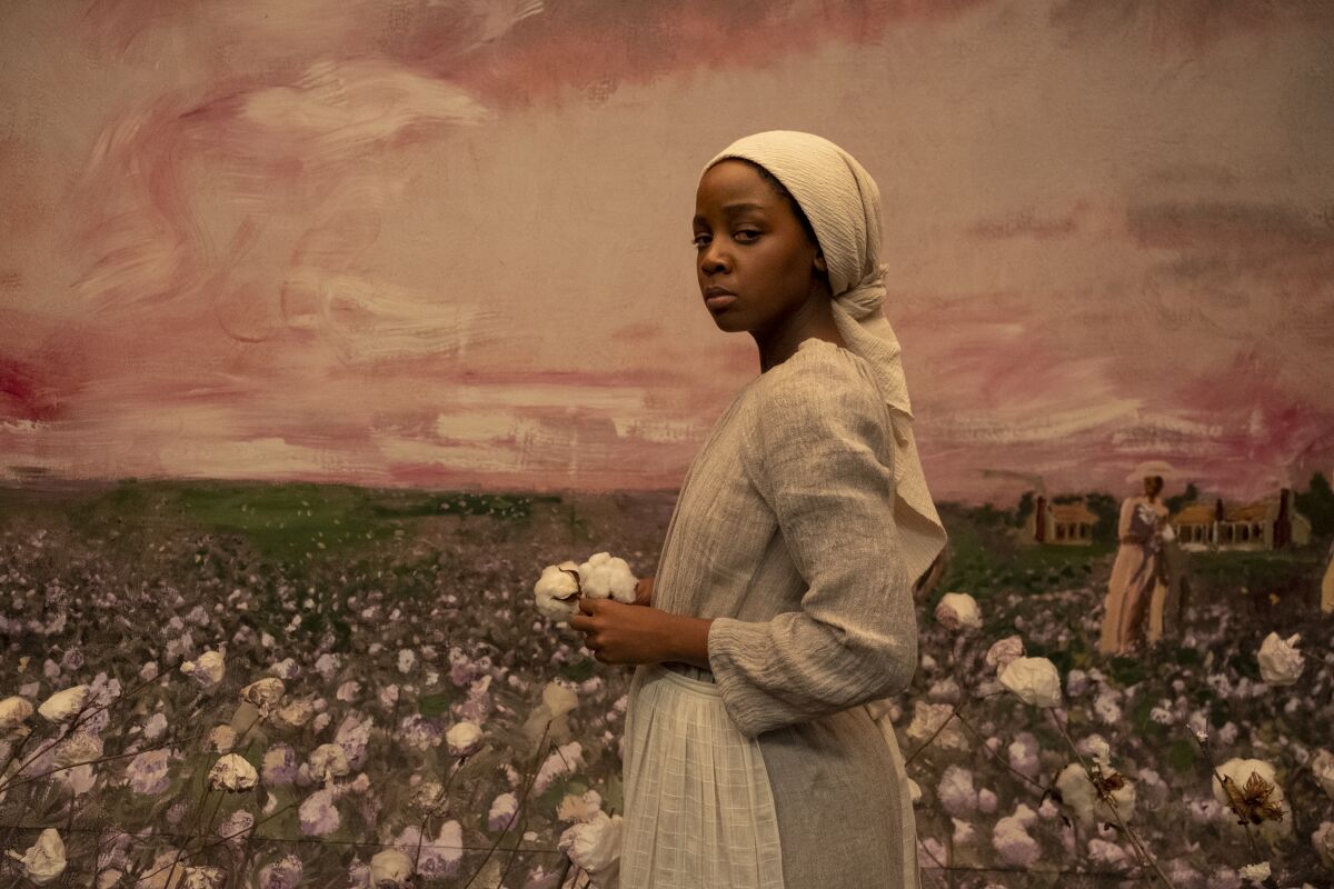 An enslaved woman looks at the camera while standing before a mural
