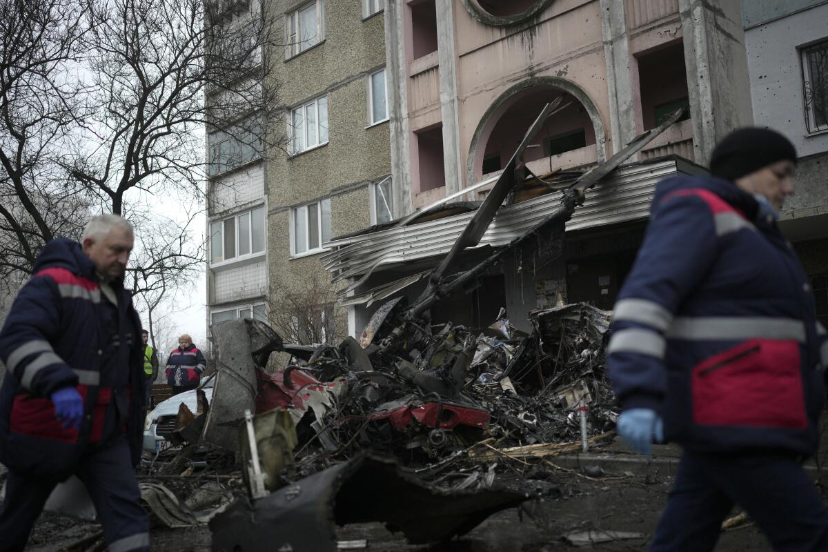 Workers in dark blue winter jackets walk past helicopter wreckage that damaged the front of a building 