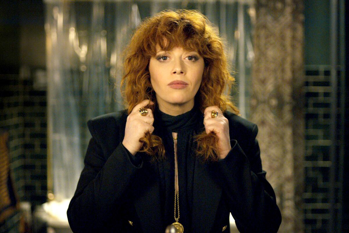 A woman wearing a black zip-up jacket adjusts her curly hair in a mirror