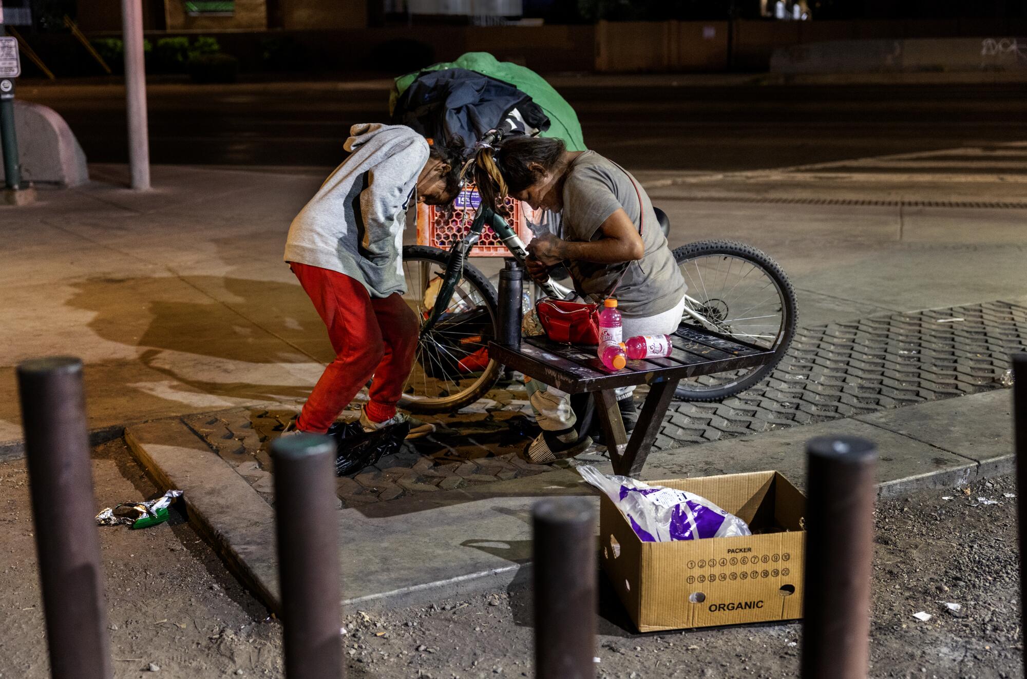 Two people next to a bike after dark, one standing hunched and one sitting, with bright-pink beverage bottles and belongings