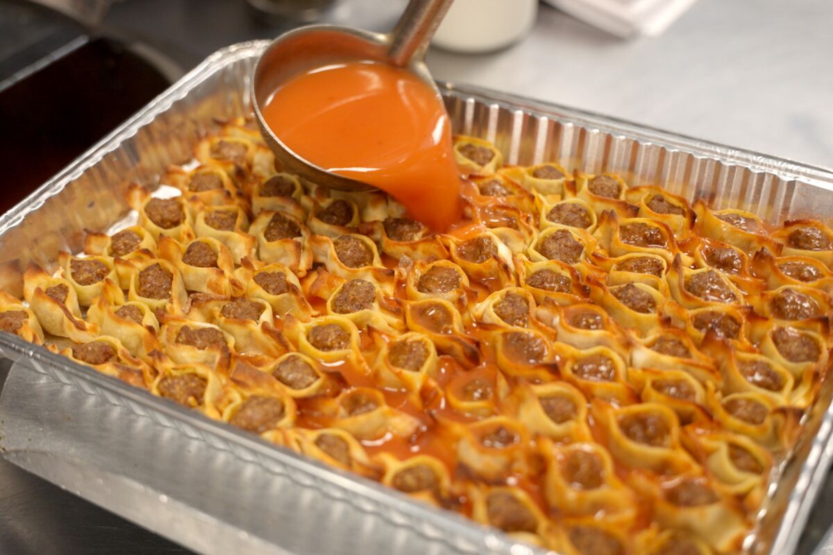 Red liquid pours from a ladle into a foil tray of pastry-wrapped meatballs