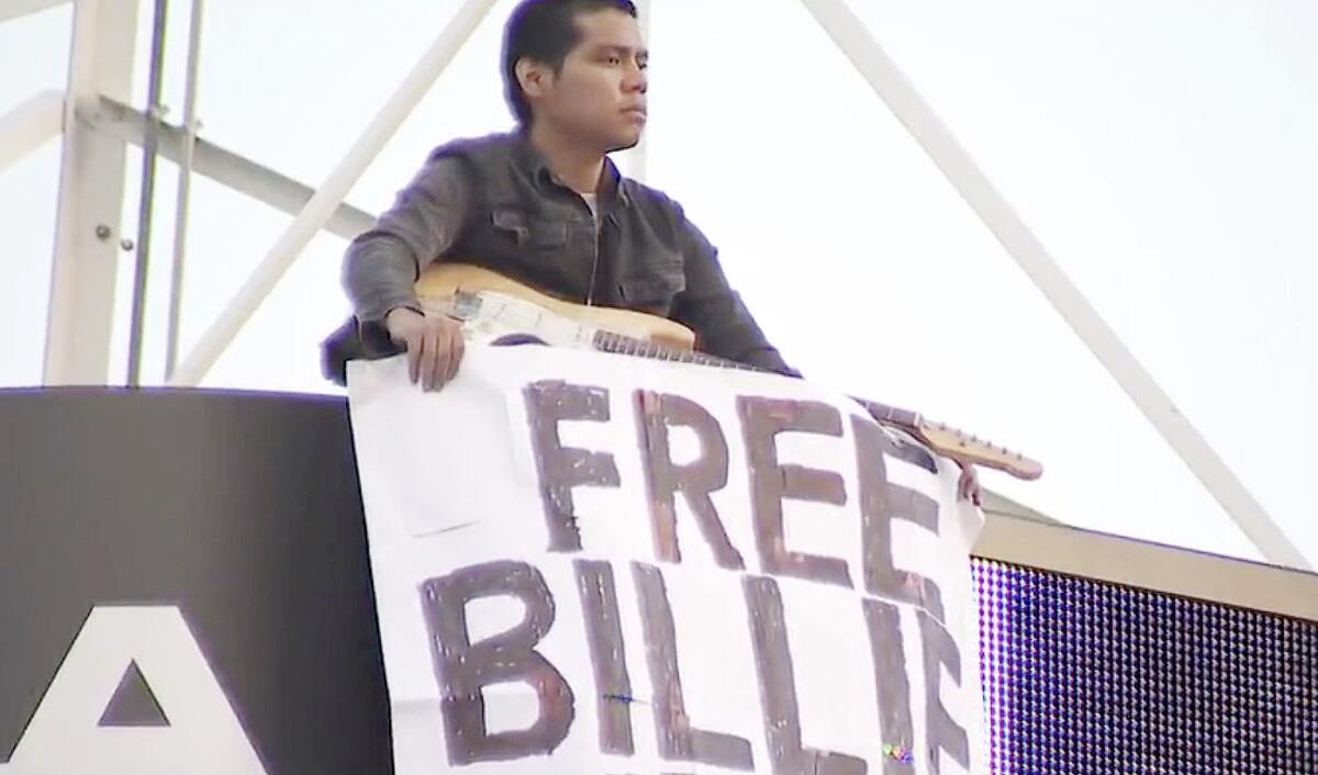 A man with a guitar and a handwritten sign with the words 'Free Billie Eilish'  stands on a TV station tower.