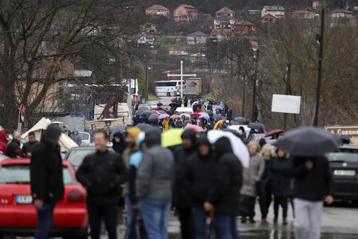 People in coats and carrying umbrellas block the road, preventing cars from moving.