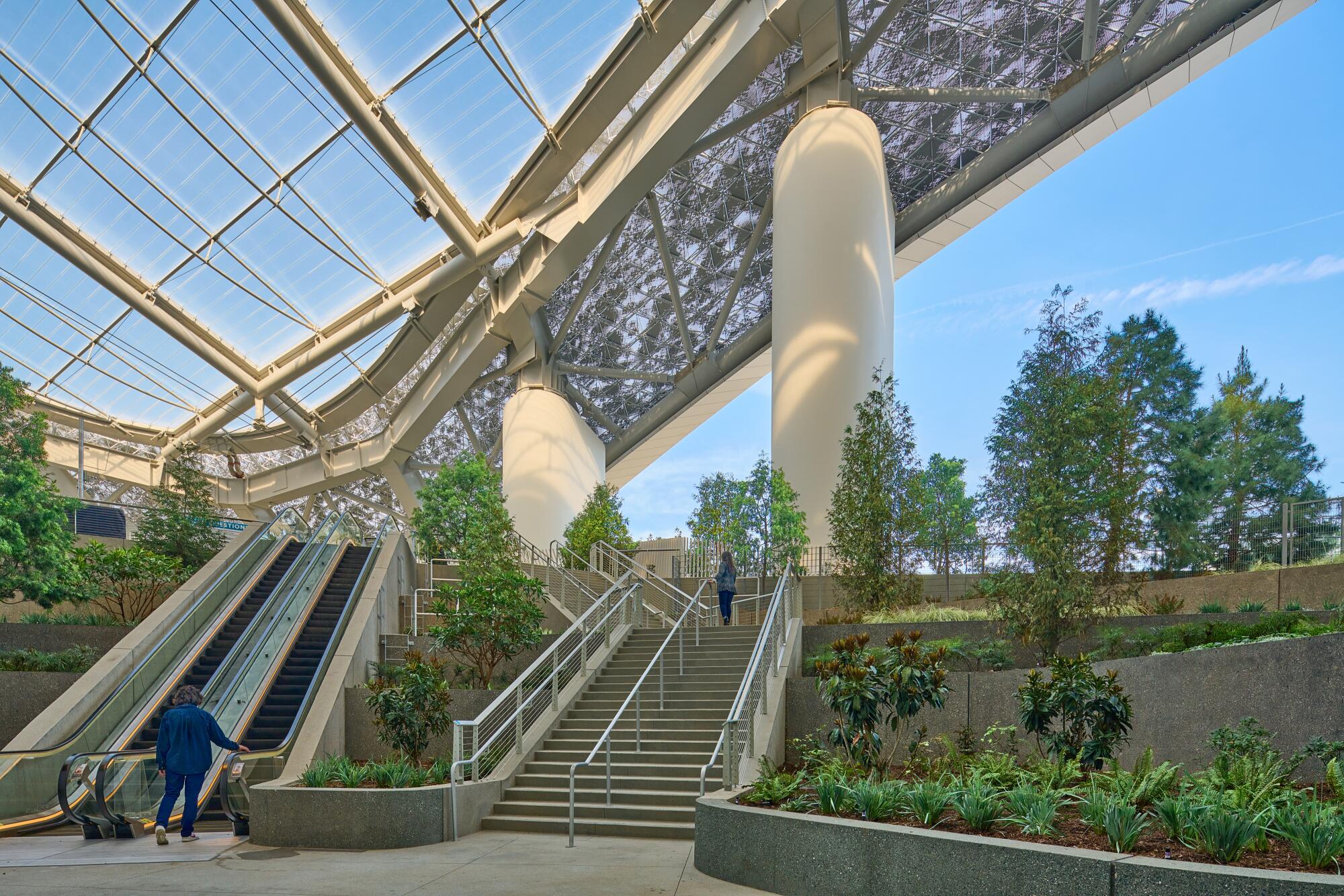 Steps and an escalator descend through terraced gardens filled with trees.