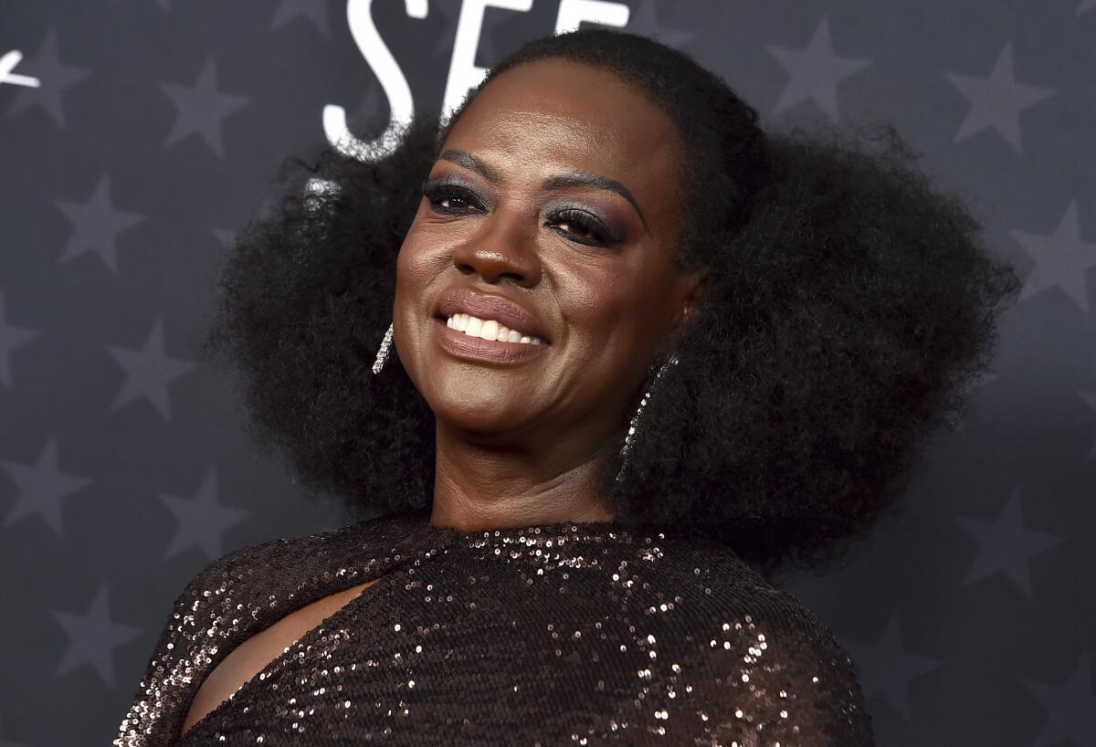 A woman in a glittering formal gown and hoop earrings smiles broadly upon arrival at an awards show