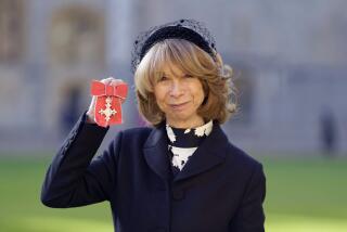 A woman in a blue hat and wool jacket and a high-neck black-and-white top holding up a red British medal of honor in one hand