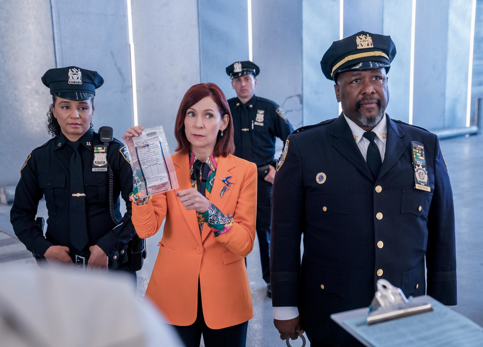 A woman in an orange blazer is flanked by a pair of officers in dark uniforms.