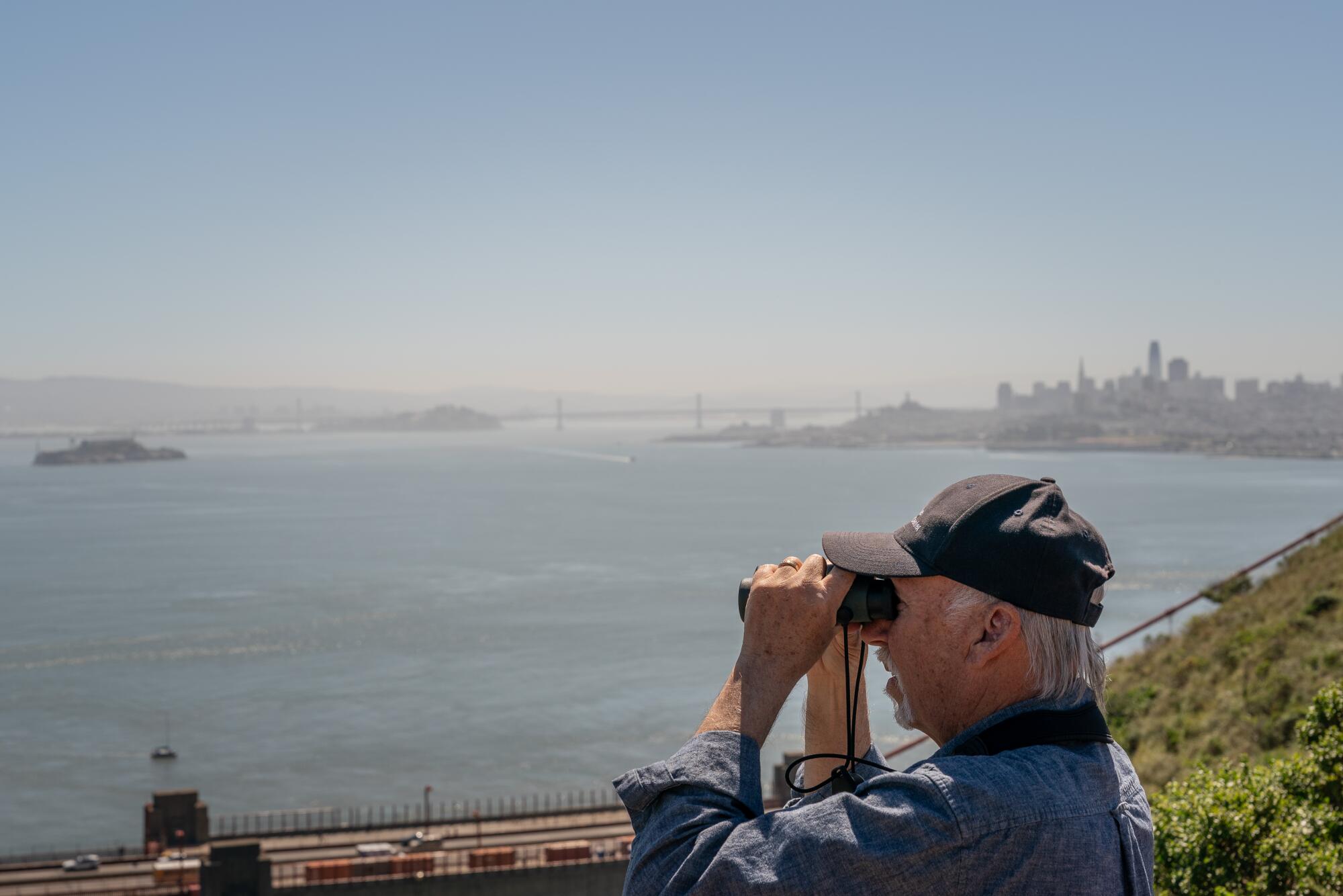 Bill Keener looks through binoculars at the San Francisco Bay, with the city skyline in the background. 