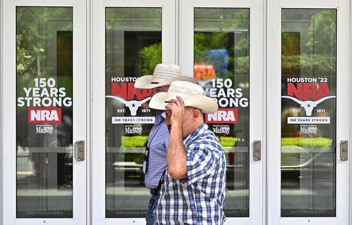 Two men in cowboy hats walking past signs reading "Houston '22 NRA" and "150 years strong" at a building's entrance.