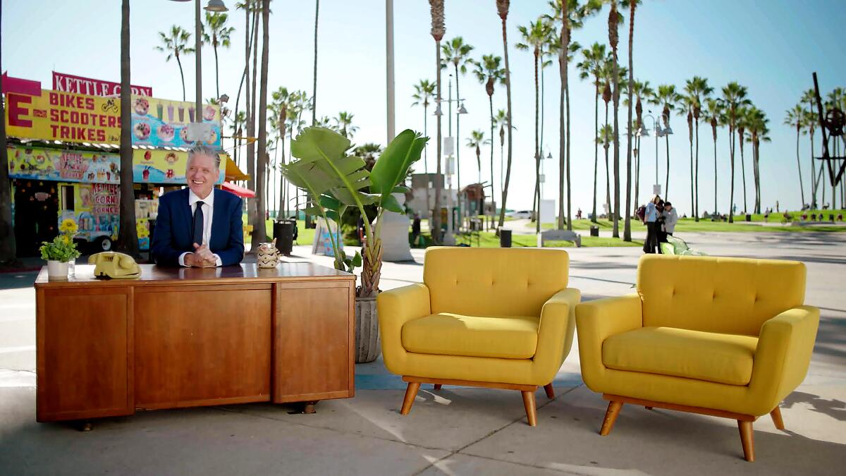 A man in a suit behind a desk next to two yellow chairs on a boardwalk.