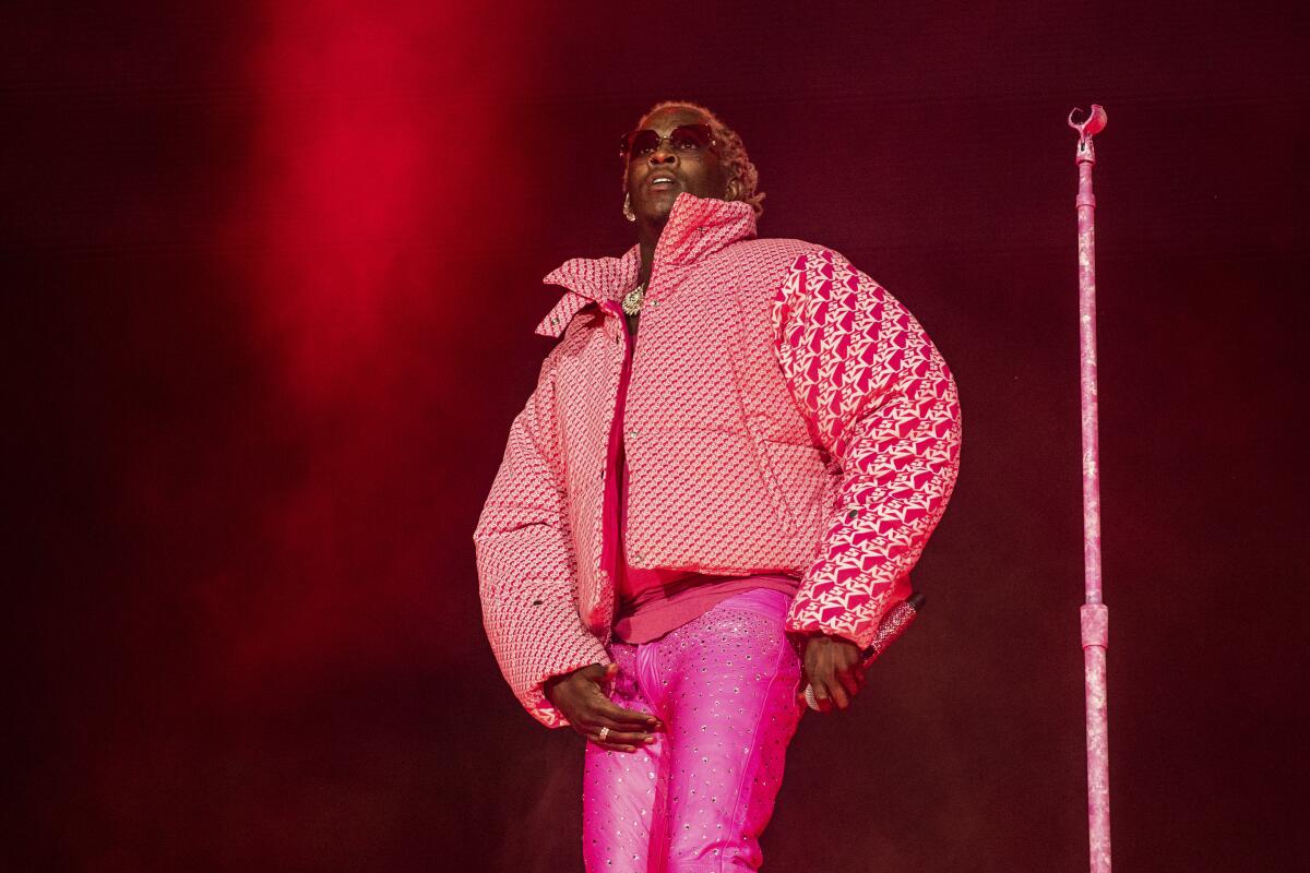A male rapper in a pink puffer jacket and hot pink pants holds a microphone down at one hip on stage
