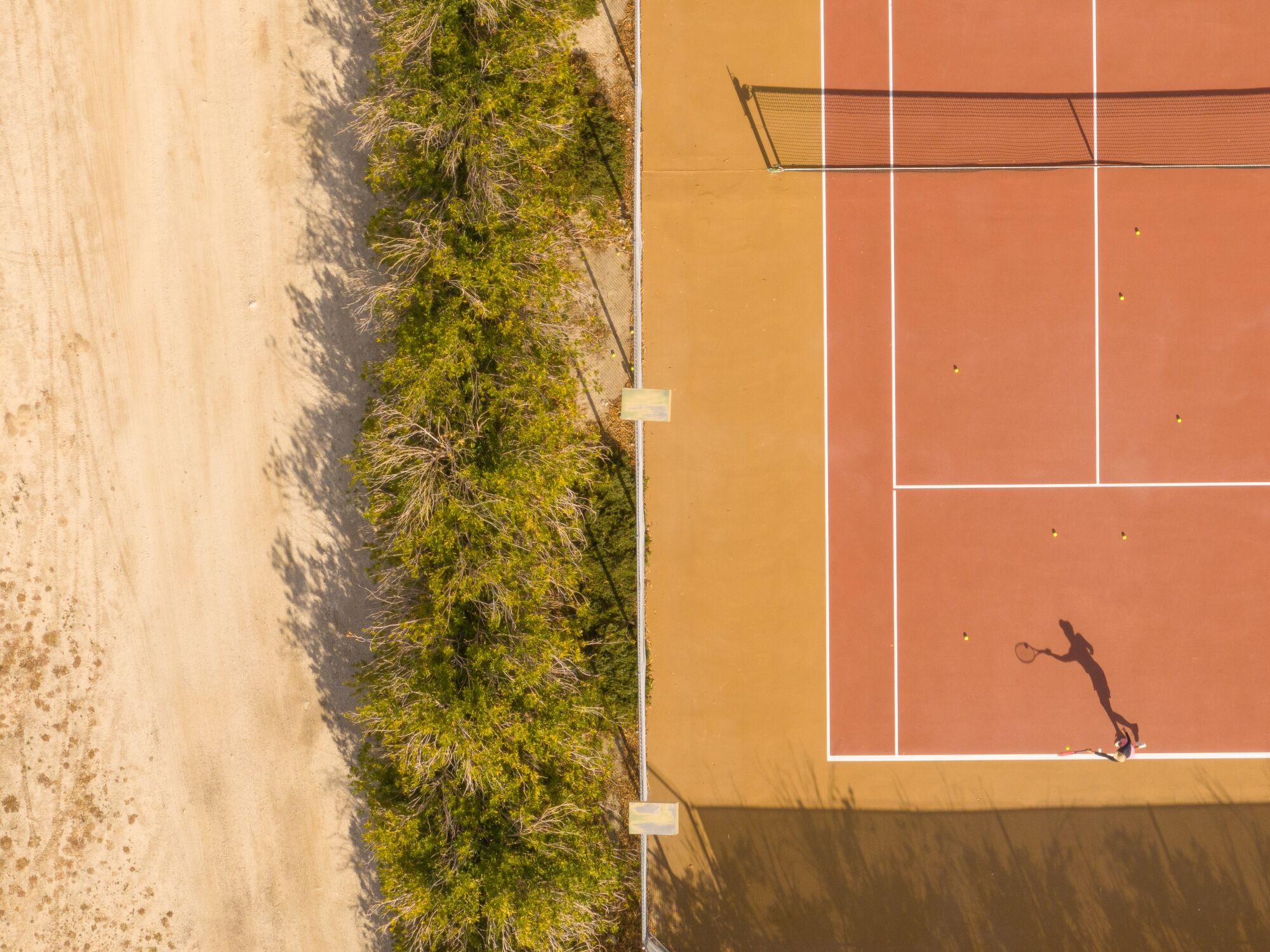 An overhead image of a tennis player and their shadow on a court that sits alongside desert sand and greenery.