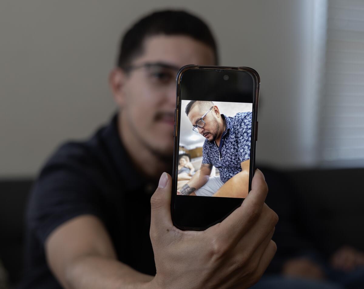 A man with dark hair and glasses holds a cellphone showing a photo of a man, also in glasses, wearing a patterned shirt