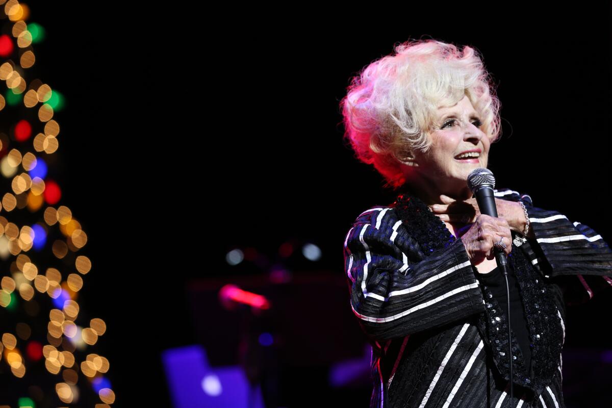 Brenda Lee wears a sequin and striped black and white ensemble as she performs in front of a Christmas tree. 