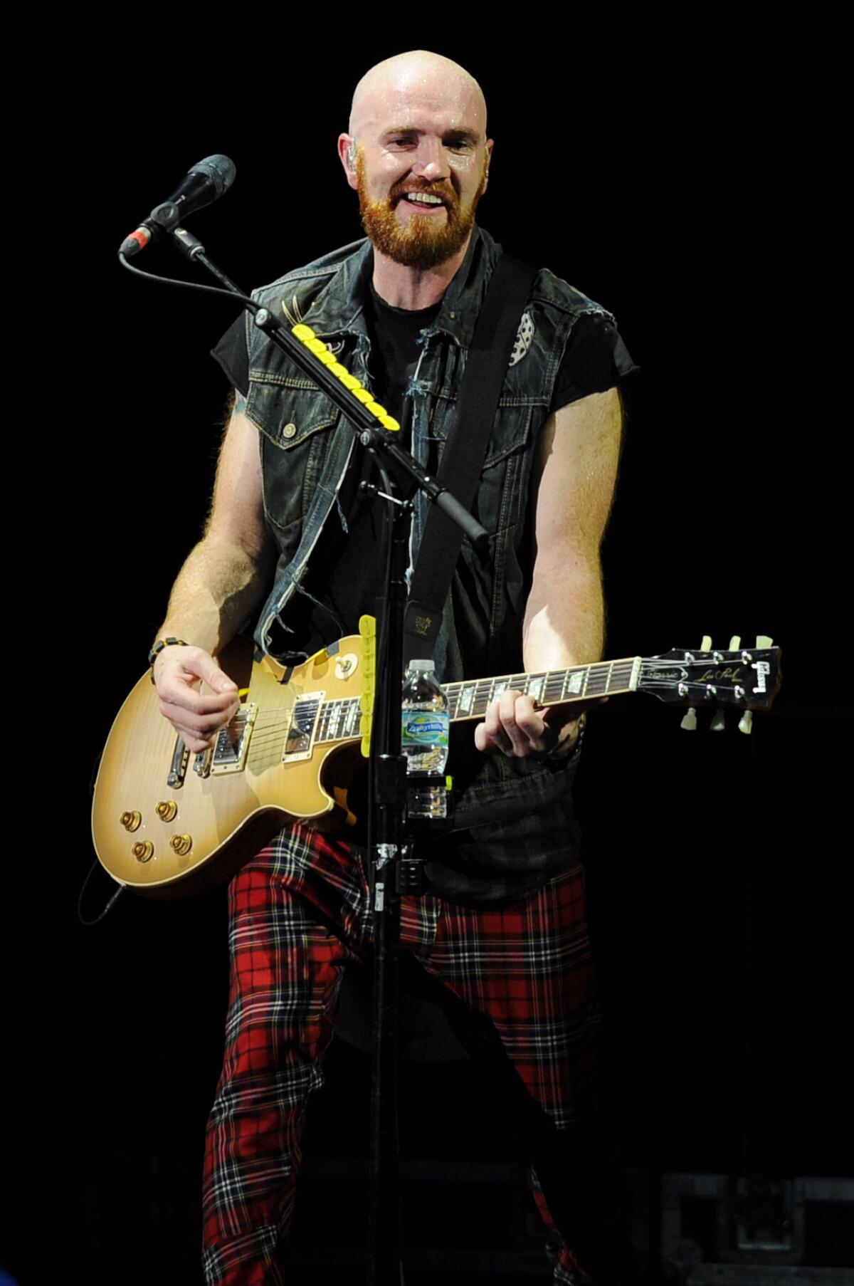 Mark Sheehan of the Script in a sleeveless denim vest holding a guitar on a stage.