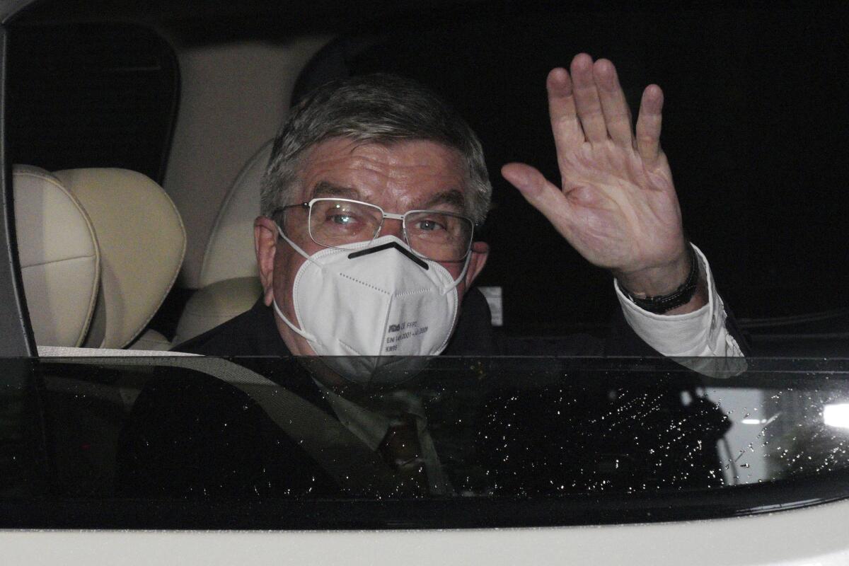 Thomas Bach, wearing a mask, waves from a vehicle.