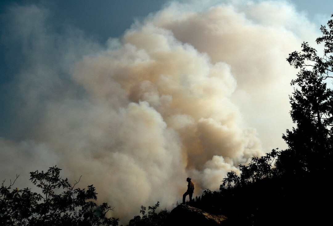 A huge plume of smoke billows skyward as a person stnads on a hill of trees