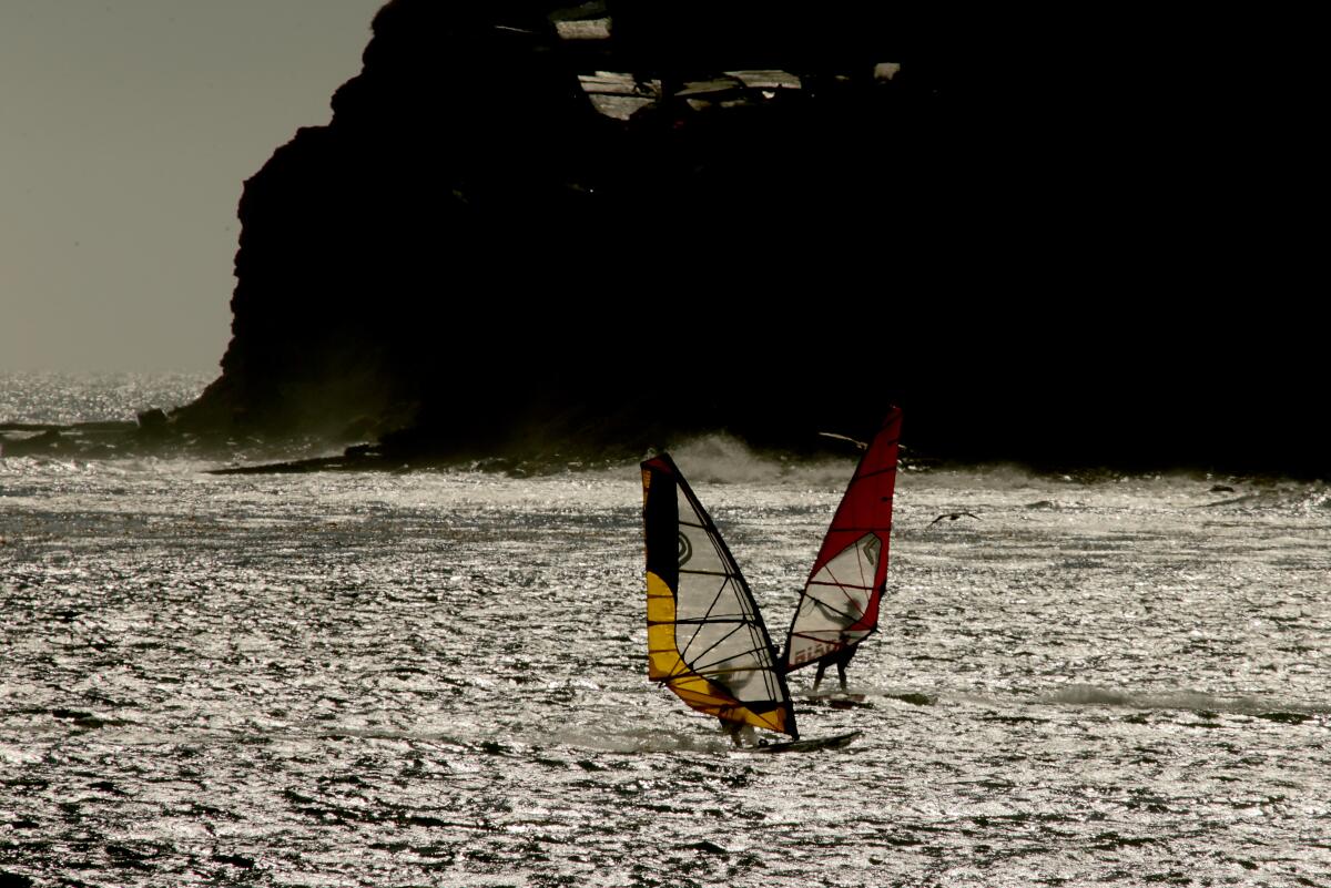 Windsurfers are shown in the ocean with a background of dark cliffs.