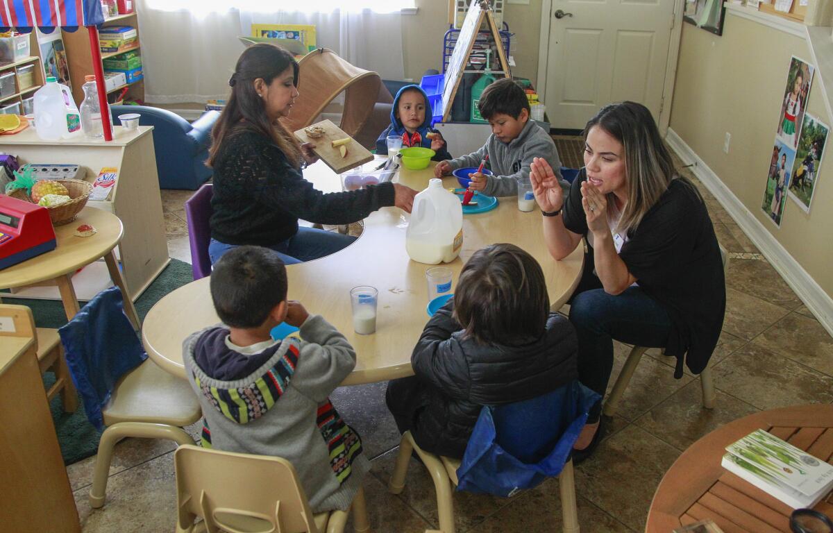 Two women sit at a table with four children eating snacks and drinking milk in a child care center