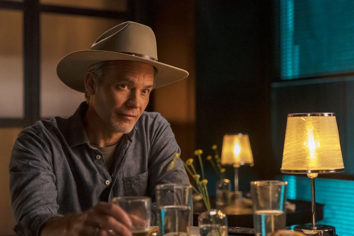 In a blue shirt and a cowboy hat, Raylan Givens sits at a table with water glasses.