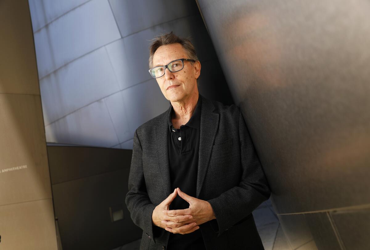 A man in a black suit stands before the stainless steel facade of Disney Hall with his hands clasped.