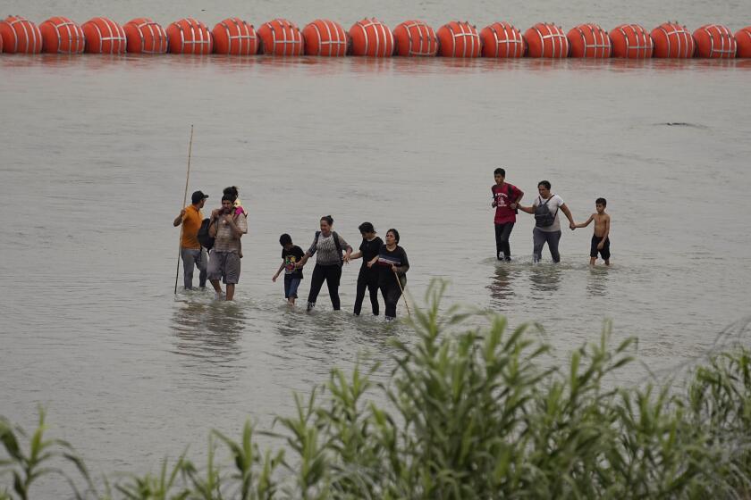 Migrants who crossed the Rio Grande from Mexico walk past large buoys being deployed as a border barrier on the river in Eagle Pass, Texas, Wednesday, July 12, 2023. The floating barrier is being deployed in an effort to block migrants from entering Texas from Mexico. (AP Photo/Eric Gay)