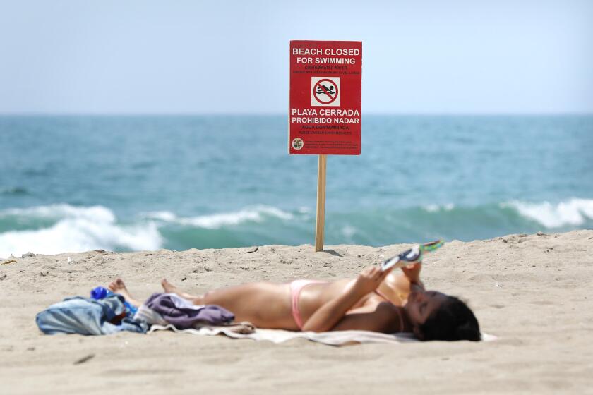 PLAYA DEL REY, CALIFORNIA: A beach goer sunbathes near a sign warning people to stay out of the water at Dockweiler State Beach on Tuesday, July 13, 2021 after a 17-million-gallon sewage spill. (Christina House / Los Angeles Times)
