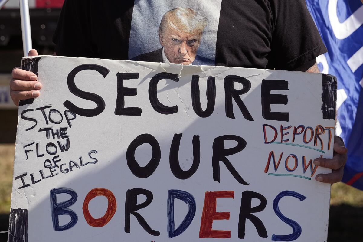 A person wearing a Trump shirt holds a handmade "Secure Our Borders" sign.
