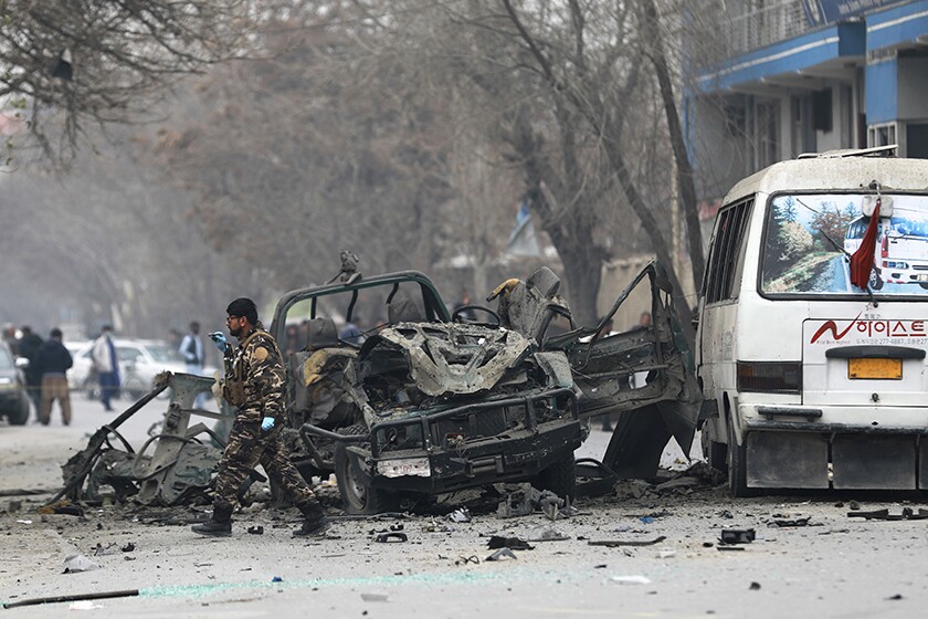 A man in camo walks next to a bombed-out vehicle on a city street in Kabul, Afghanistan. 