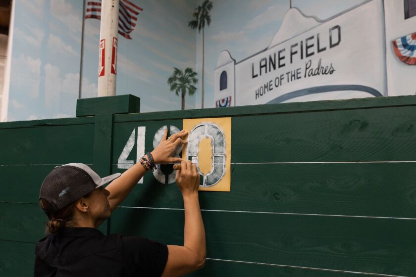 Local muralist Hanna Daly uses a stencil and brush to paint Lane Field’s original distance (480 feet) from home plate to center field, at Glenner Town Square’s new Wiffle ball court in Chula Vista on Tuesday, March 21, 2023. The downsized court’s distance from home plate to center field is 35 feet, making it approximately a 1/15 scale model of Lane Field, which was the original home for the Padres. Glenner Town Square is an adult day care center that provides reminiscence therapy for seniors with dementia.