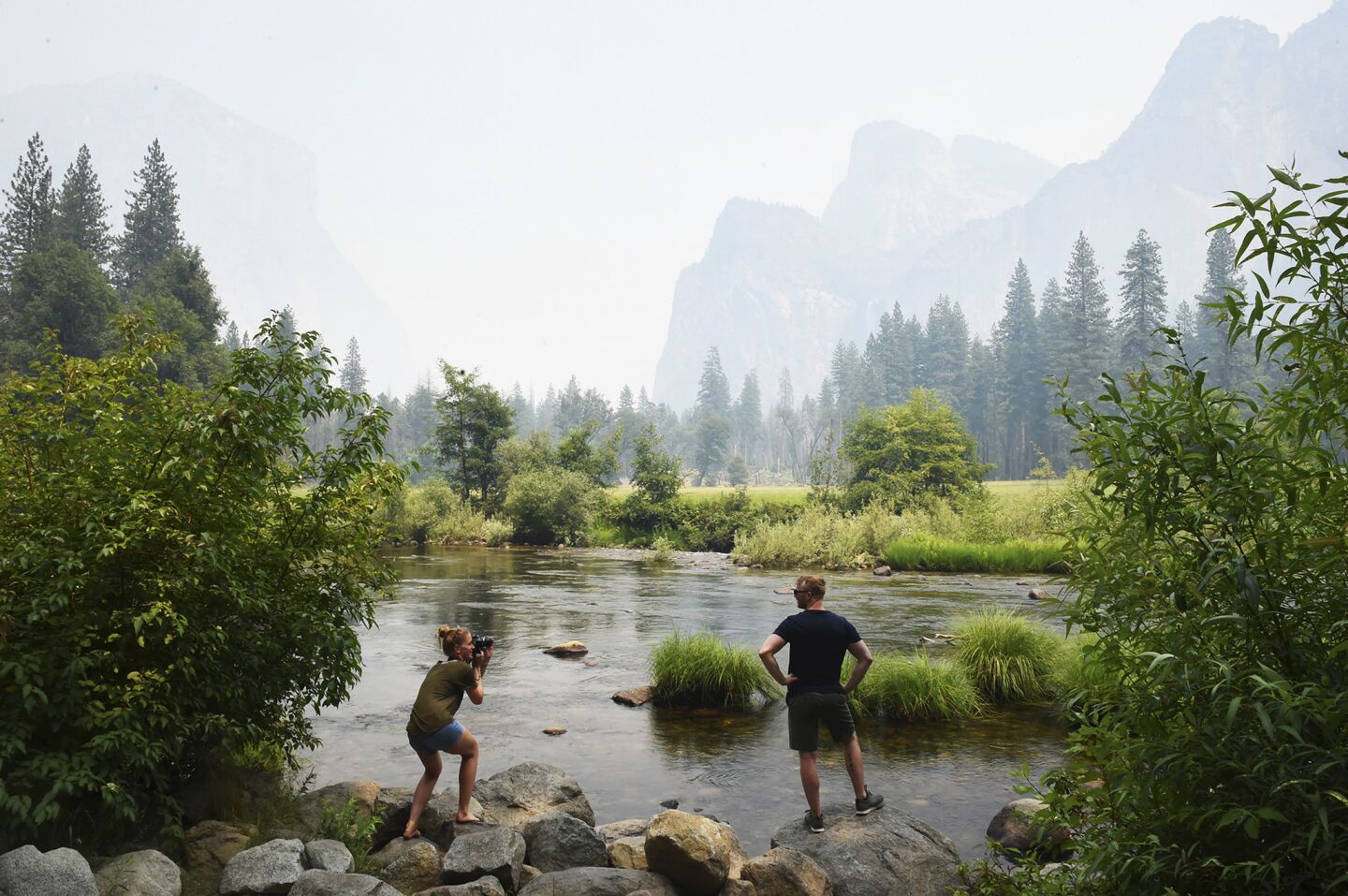 German tourist Stephanie Schultz, left, photographs Kai Rudolph along the Merced River in Yosemite Valley as smoke from the Ferguson fire hangs in the air in Yosemite National Park.