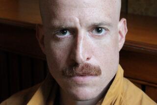 A man with shaved head and red mustache in a yellow shirt, staring intensely.