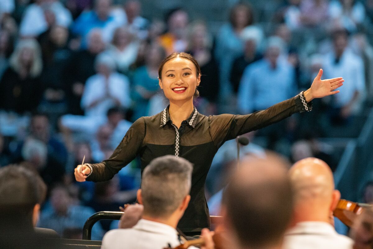 Tianyi Lu holds a conductor's baton while extending an open arm to a group of seated musicians 