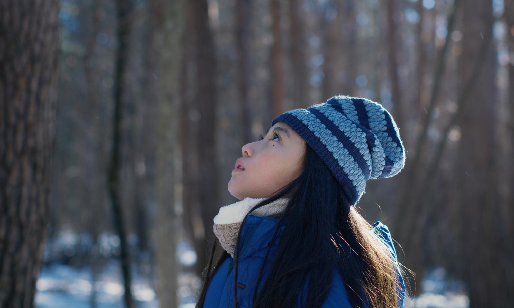A girl in the woods wearing a knit hat looks up.