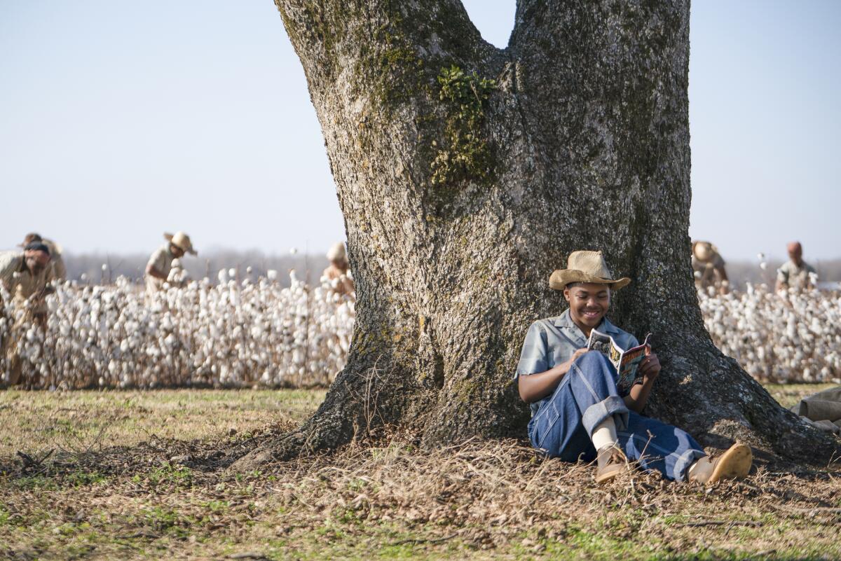 A boy sits at the foot of a tree reading a book.