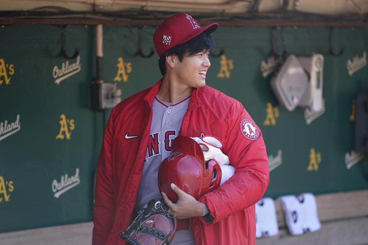 Shohei Ohtani stands in the dugout, smiling and holding his batting helmet and glove.