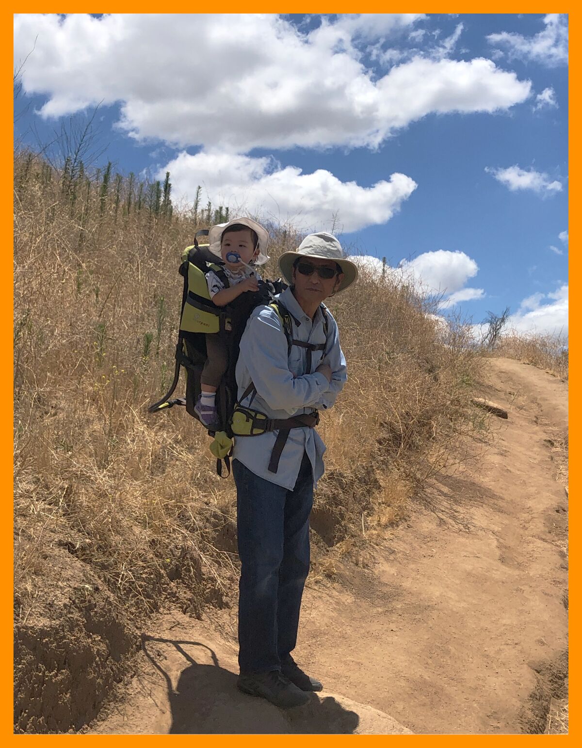 A man in a hat stands with a baby in a carrier on his back on a dirt path under a blue sky with puffy white clouds.