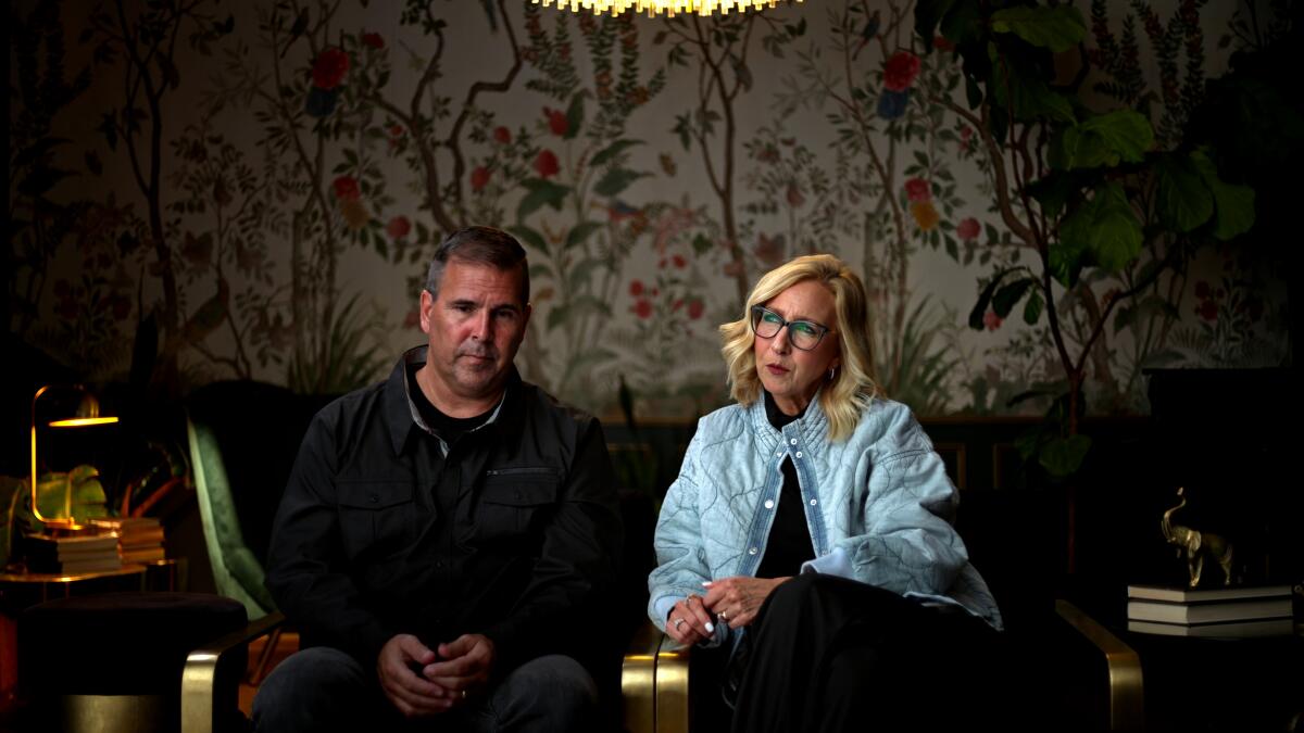 Dean and Kelly Wilking sit side by side in a dim room with ornate floral wallpaper