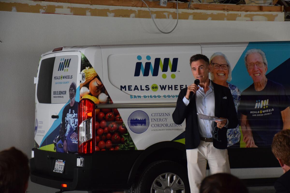 a man holds a microphone while speaking in front of a Meals on Wheels delivery van 