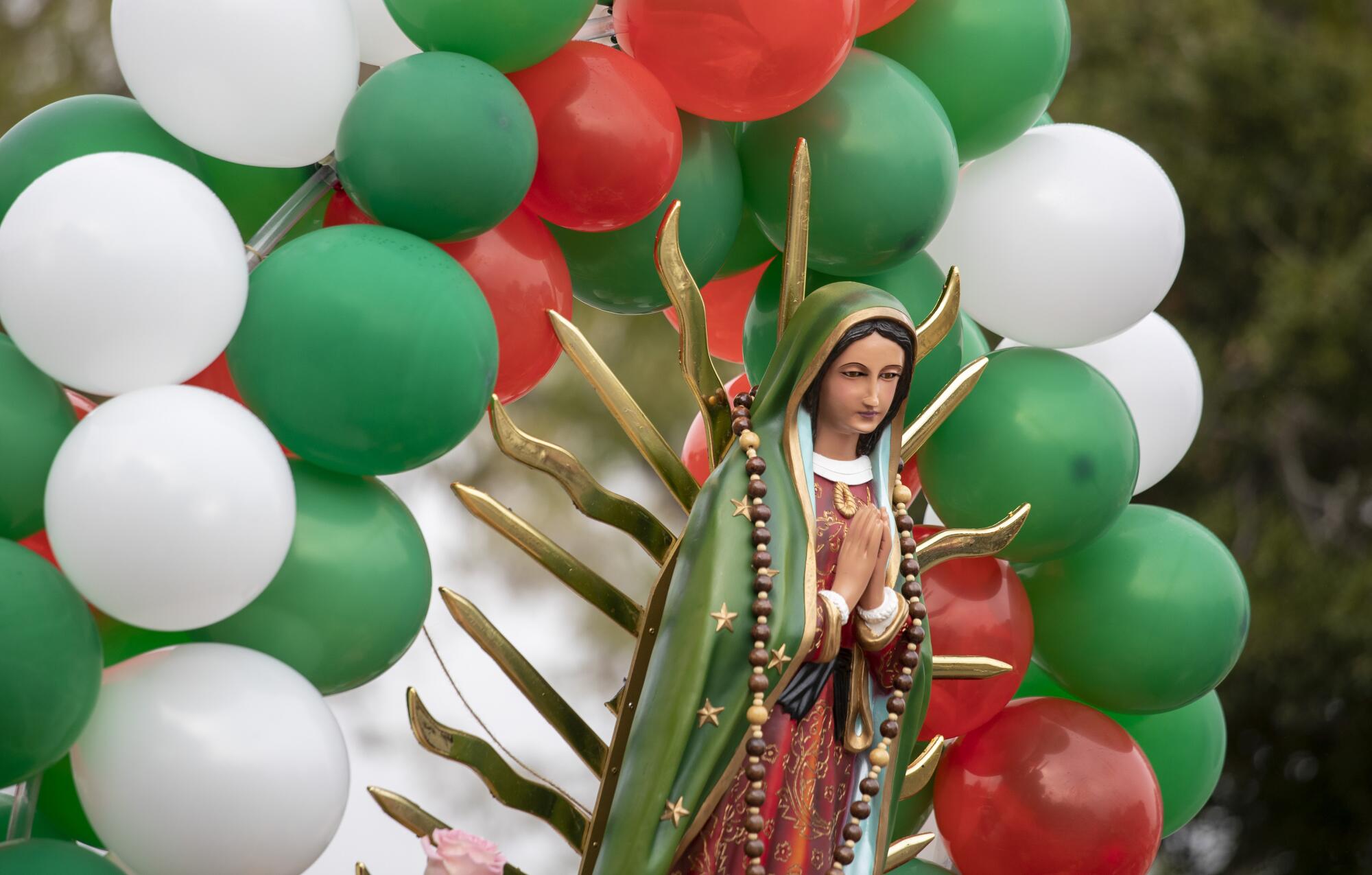 Rays of gold emanate from a statue of the Virgin of Guadalupe, surrounded by red, green and white balloons