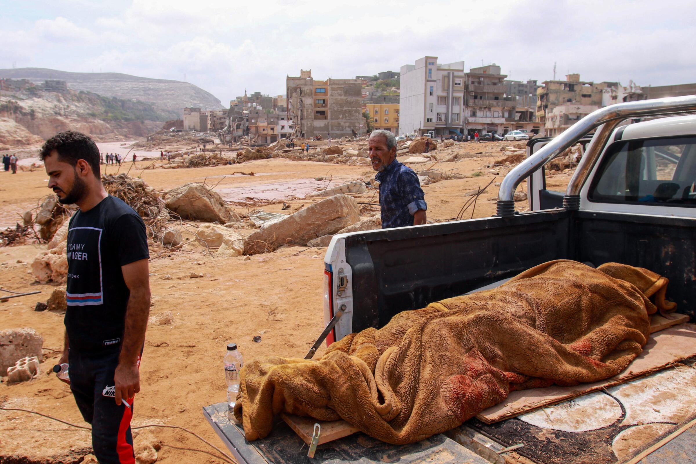 Two men stand near a body wrapped in a brown blanket on a pickup truck bed 