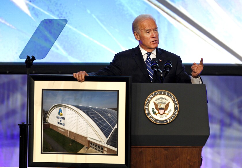 Joe Biden speaks at a podium alongside a framed photo of a building.