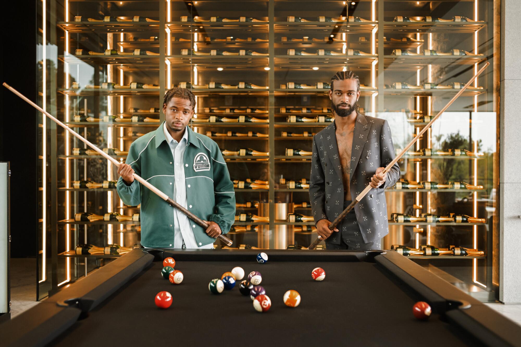 Two men stand wearing Louis Vuitton stand at the head of a pool table holding cues.